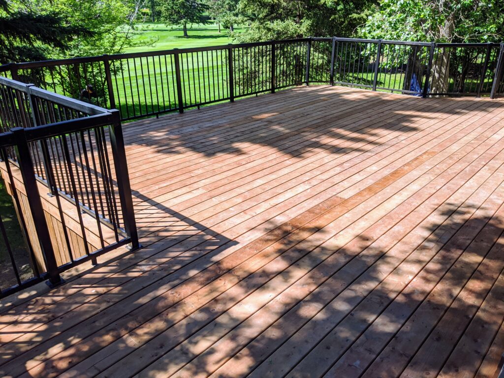Sunlit wooden deck with black railing overlooking a green yard.
